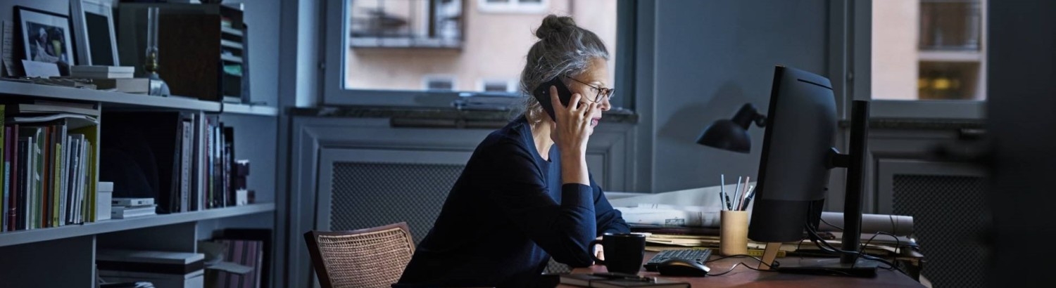 Kvinna vid skrivbord i hemmamiljö, pratar i telefon framför dator. 