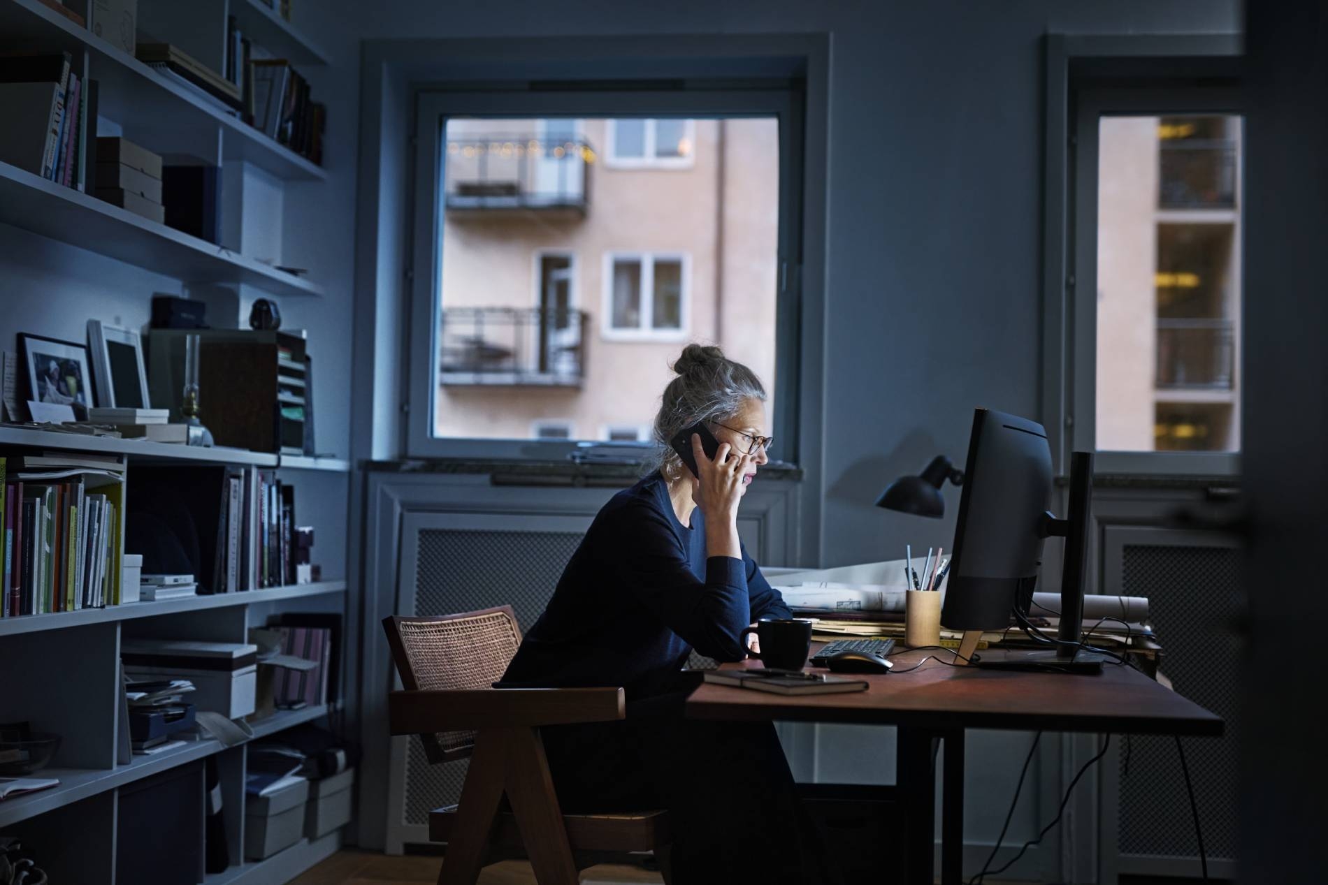 Kvinna vid skrivbord i hemmamiljö, pratar i telefon framför dator. 
